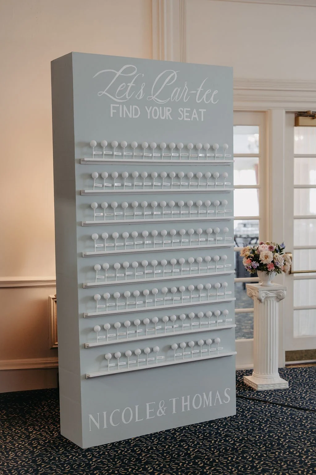 A white seating chart display with multiple labeled rows of white ball seats, titled "Let's Eat-ee" and "Find Your Seat." It has the names 'Nicole & Thomas' at the bottom and is placed in a decorated venue with floral arrangements and large window do