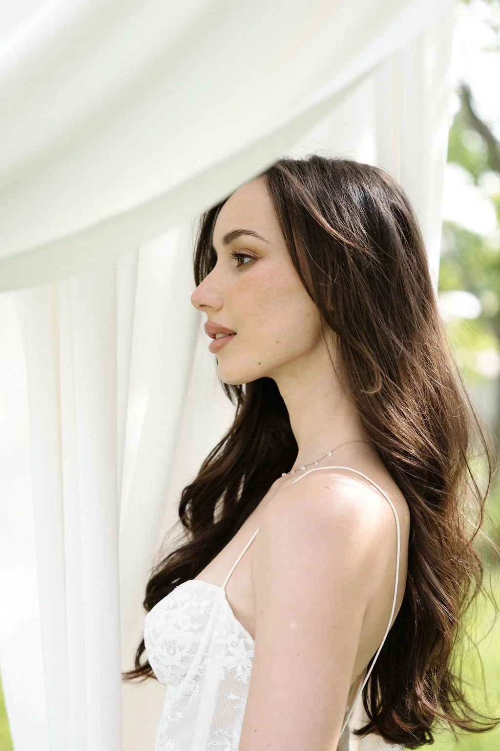 Profile of a young woman with long brown hair, wearing a white lace top and a delicate necklace, standing outdoors near white curtains with greenery in the background.