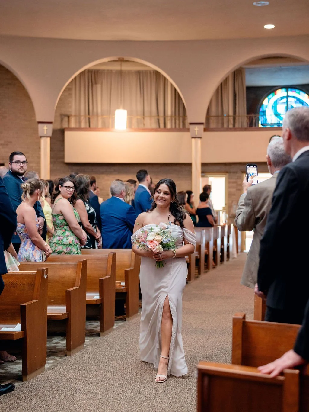A bride in an off-shoulder white dress holding a bouquet of flowers, walking down the aisle in a church, with guests watching and taking photos.