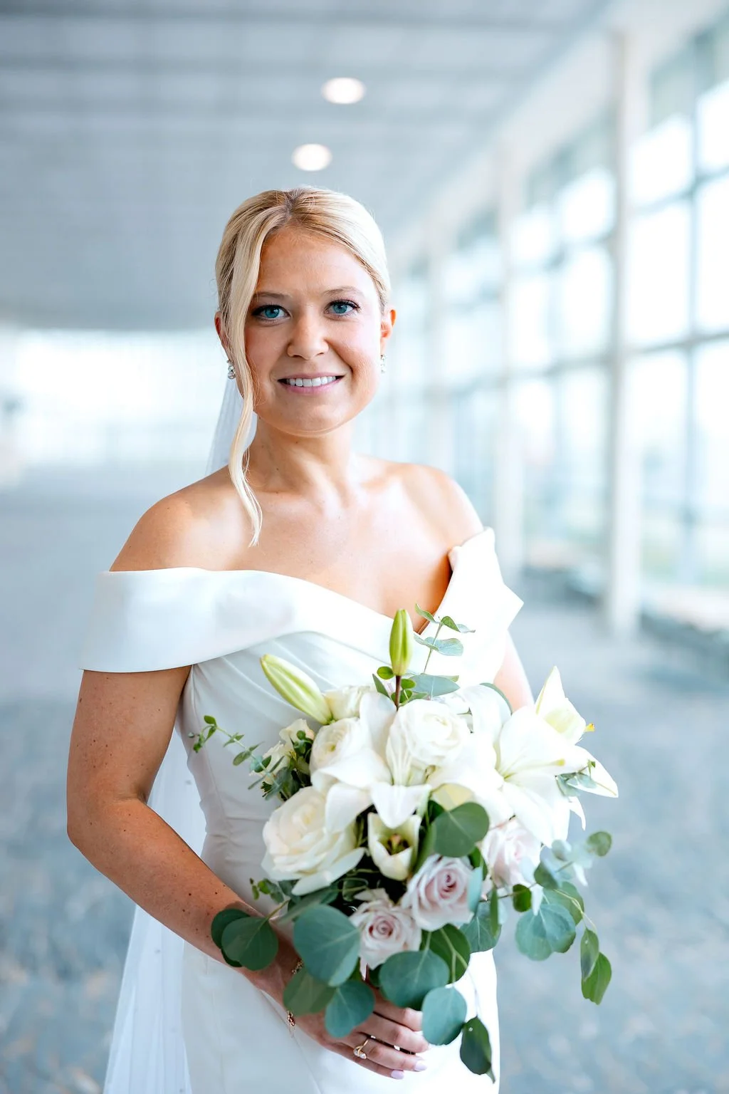 A bride holding a bouquet of white flowers and greenery inside a modern building with large windows.
