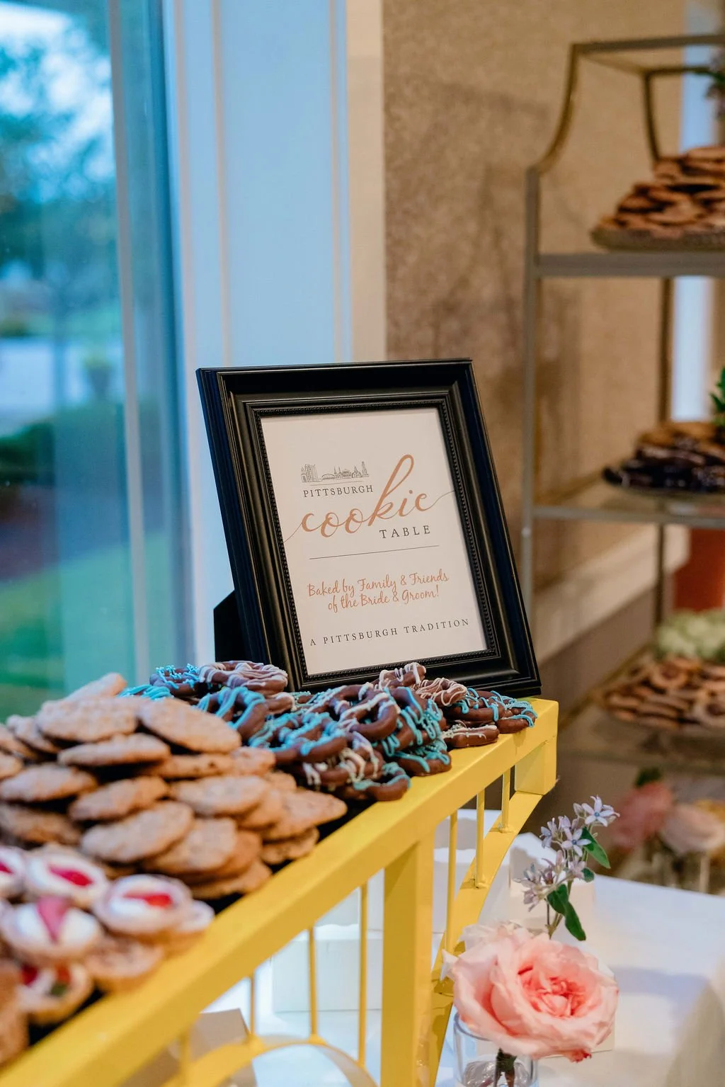 A display of cookies on a yellow stand with a framed sign that reads 'Pittsburgh cookie table' and mentions baked by family and friends for a wedding. There is a pink flower in a small vase nearby, and a shelf with more cookies in the background.
