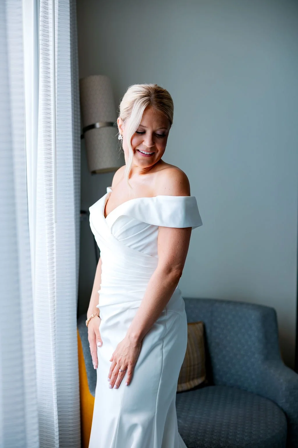 A smiling woman in a white off-shoulder wedding dress standing near a window with natural light coming in.