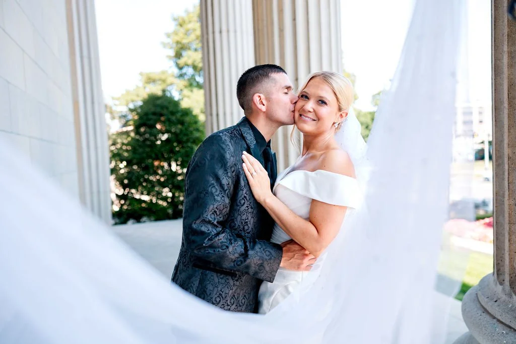 A newlywed couple, the groom kissing the bride on her cheek, standing between white columns outdoors with greenery in the background, celebrating their wedding day.