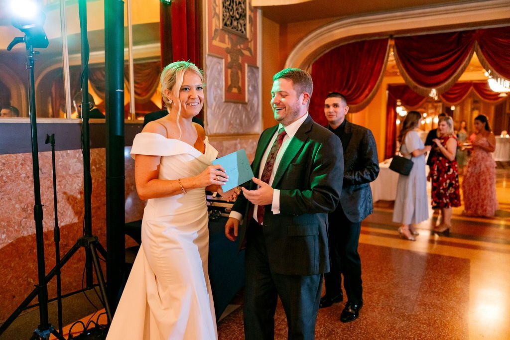 A woman in a white wedding dress is holding a blue gift bag and smiling at a man in a black suit who is giving her a present at a wedding reception. Other people are socializing in the background inside an ornate hall with red curtains and chandelier