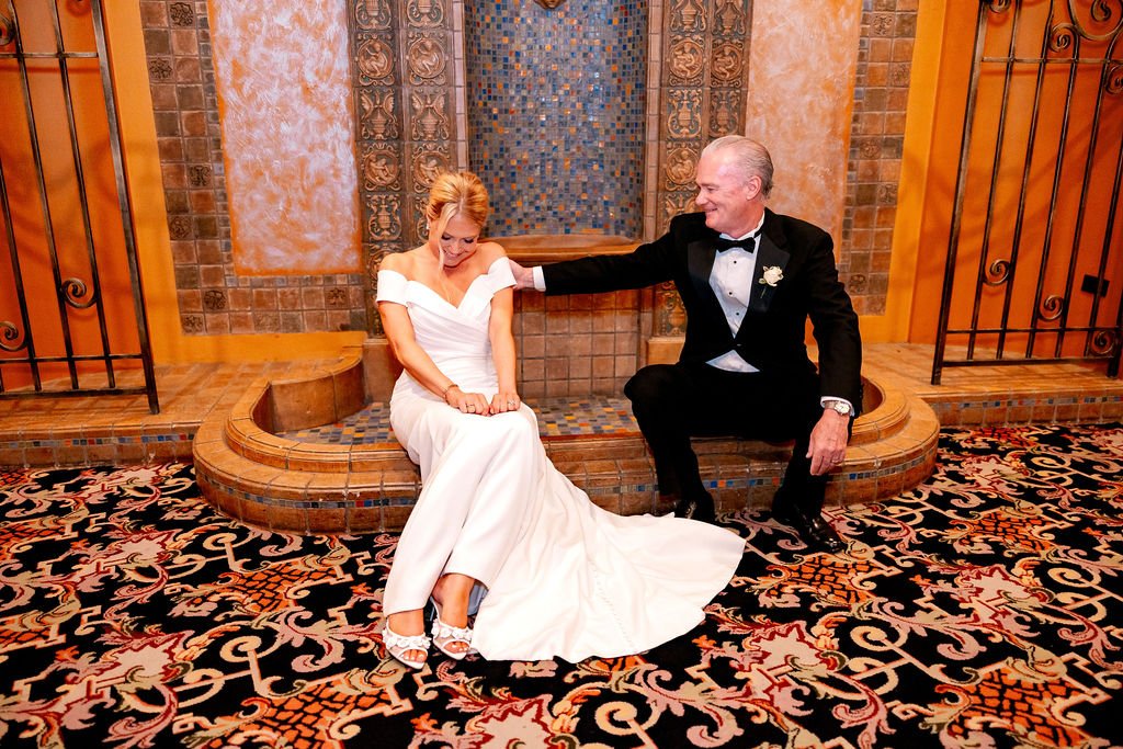 A bride and groom in wedding attire sitting in front of a decorative fountain, with the groom reaching out to touch the bride's shoulder.