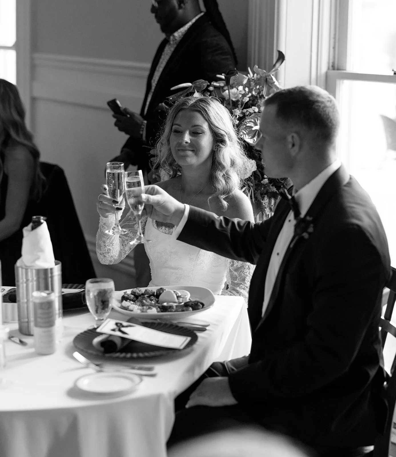In this black and white photo, a bride and groom are raising glasses in a toast during their wedding reception. The bride is smiling and wearing a lace dress, while the groom is in a tuxedo. There is a table with food and drinks in front of them and 