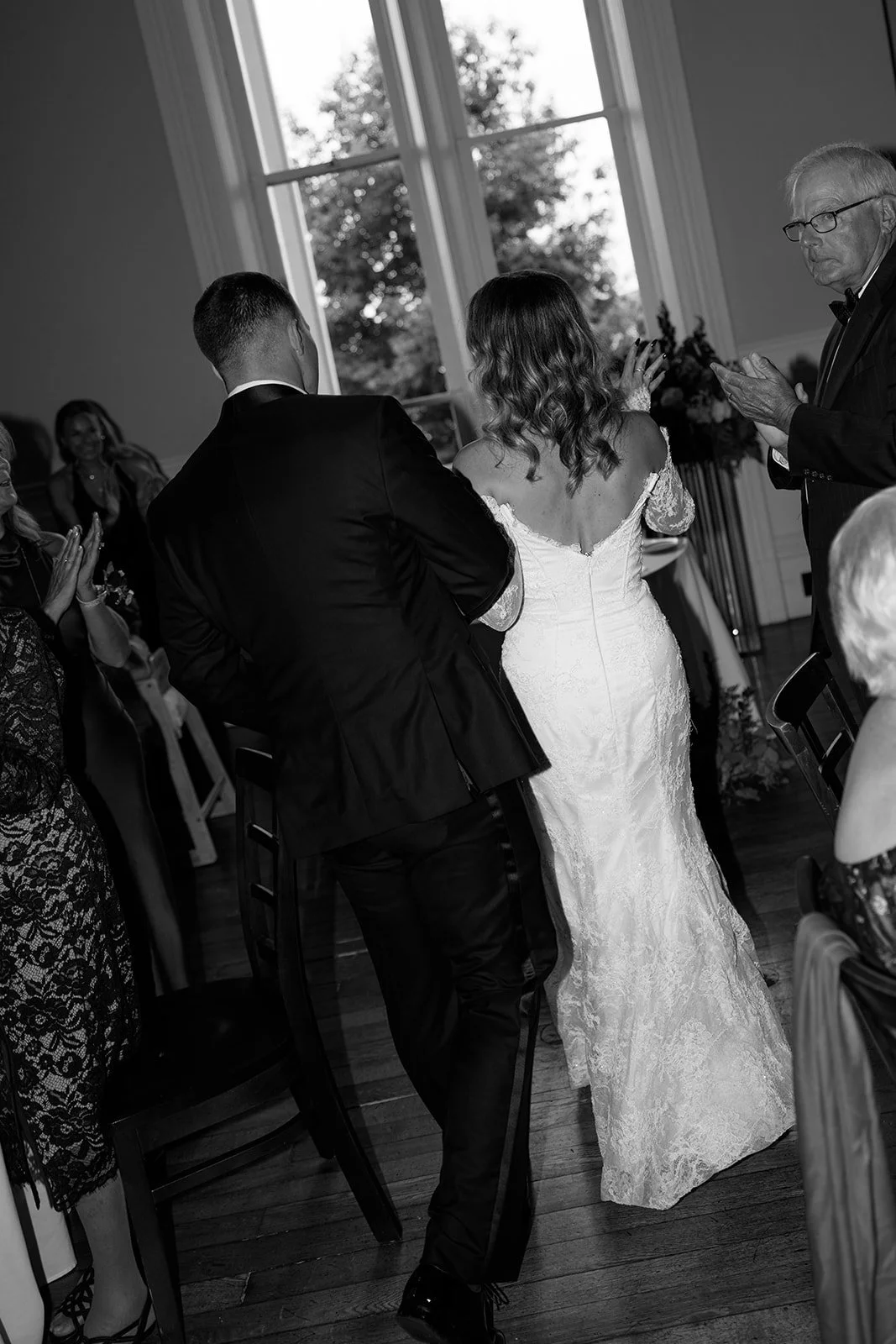 Black and white photo of a wedding reception with a bride in a lace wedding dress and a groom in a tuxedo dancing surrounded by seated guests, with a large window in the background.