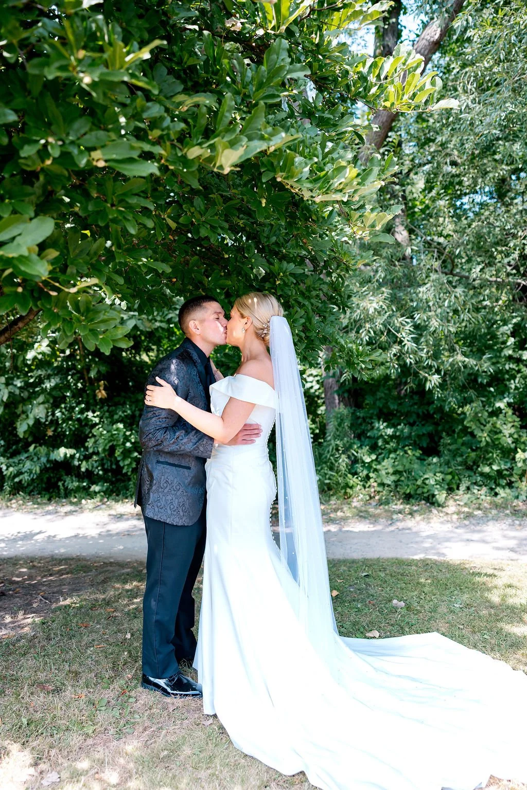 A bride and groom kissing outdoors under a large leafy green tree, with the bride wearing a white wedding gown and veil, and the groom in a black suit with a patterned jacket.