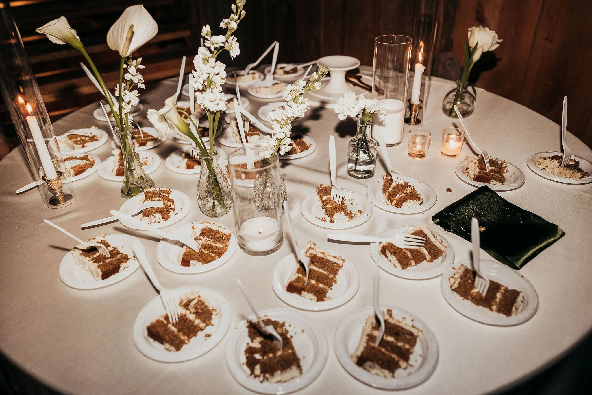 A table set for a celebration with slices of chocolate cake on small plates, floral centerpieces with white flowers, candles in tall glass holders and small votive candles, and some empty space on the table.