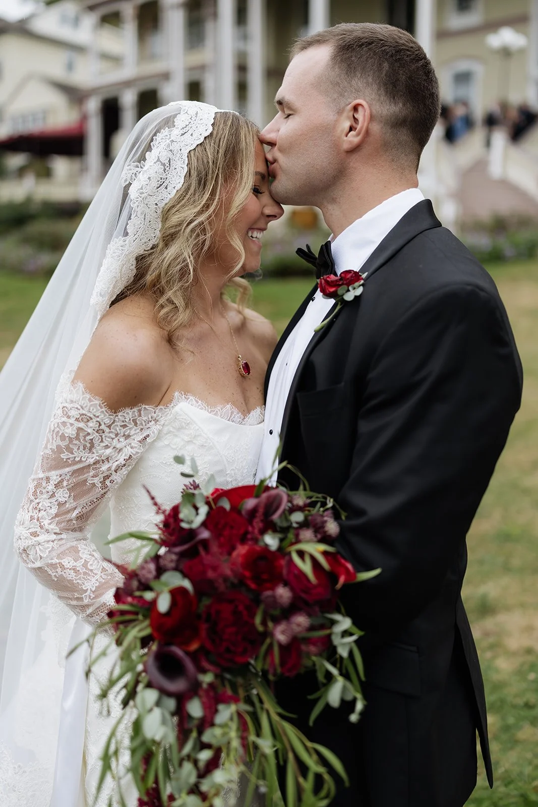 A bride and groom share a tender moment outdoors, with the groom giving a gentle kiss on the bride's forehead. The bride wears a white lace wedding dress and veil, and holds a large red and burgundy bouquet. The groom is dressed in a black tuxedo wit