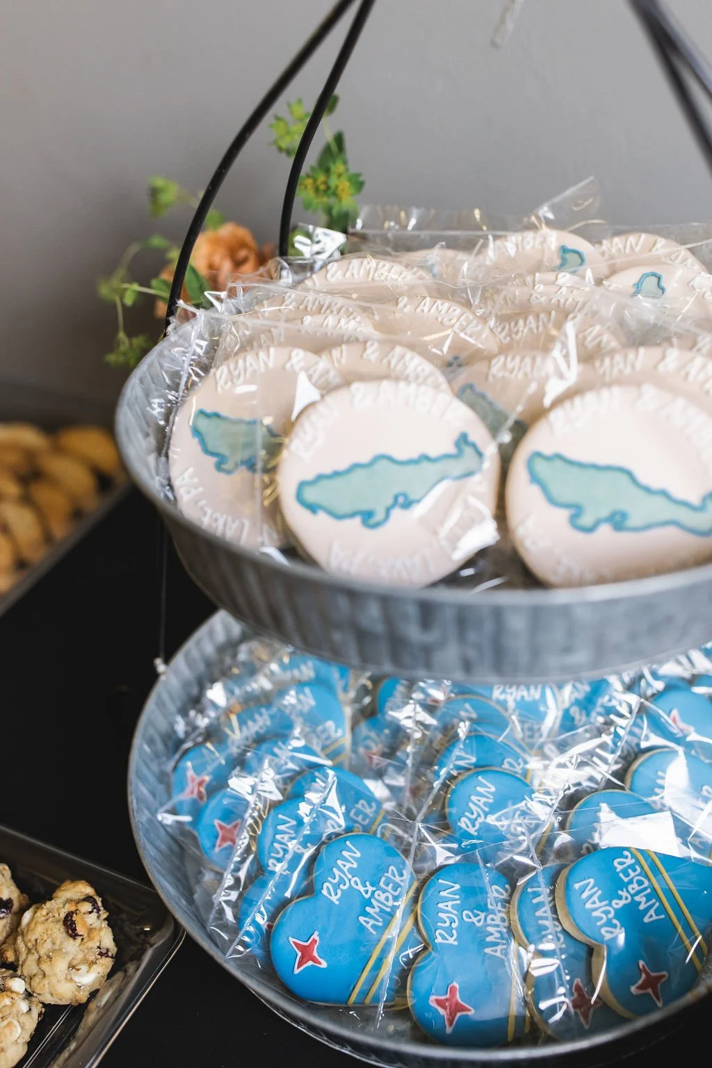 Cookies with blue and white icing featuring an island outline, branded with 'Ryan & Amber', displayed on a two-tiered metal stand at a celebration or event.