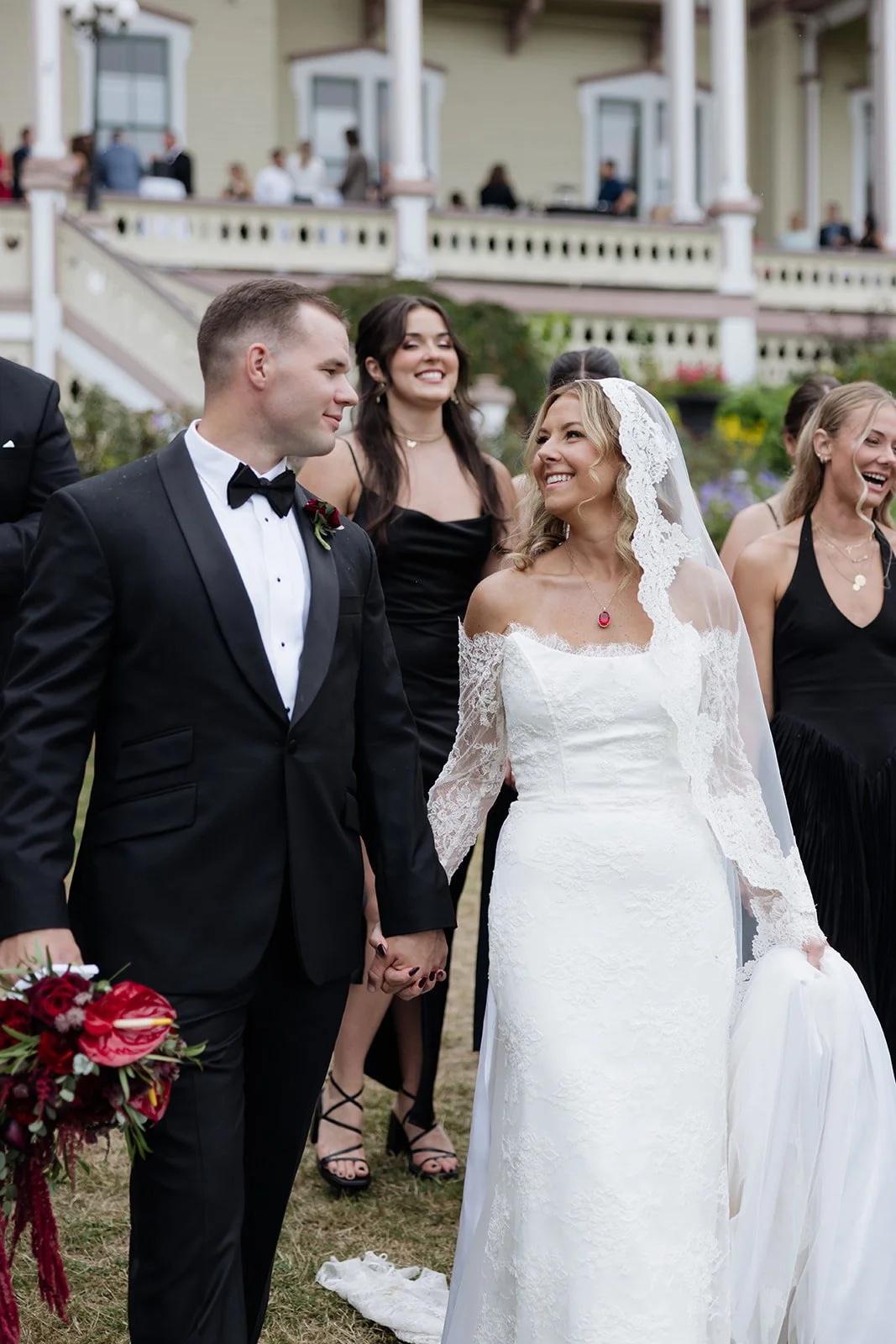 A bride and groom holding hands, smiling at each other during their wedding ceremony outdoor. The bride is wearing a white lace wedding dress and veil, the groom is in a black tuxedo with a bow tie. Bridesmaids in black dresses stand behind them, smi
