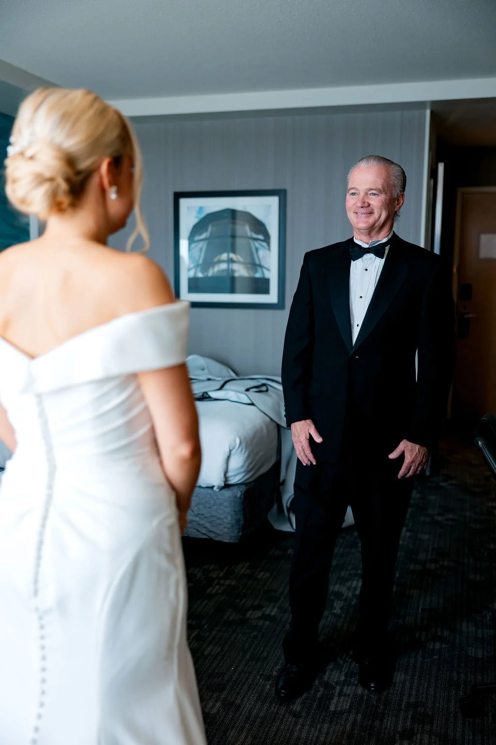 A young woman in a white off-shoulder wedding dress standing in front of an older man in a tuxedo, smiling inside a hotel room.