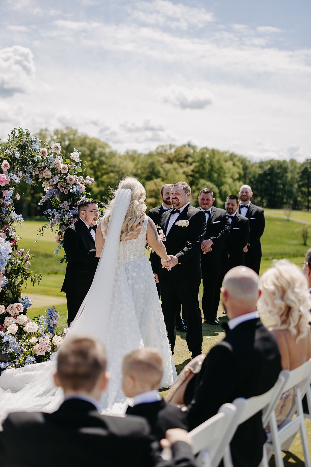 A bride and groom hold hands and look at each other during their wedding ceremony outdoors on a sunny day with a blue sky and green trees in the background, surrounded by their wedding party and guests.