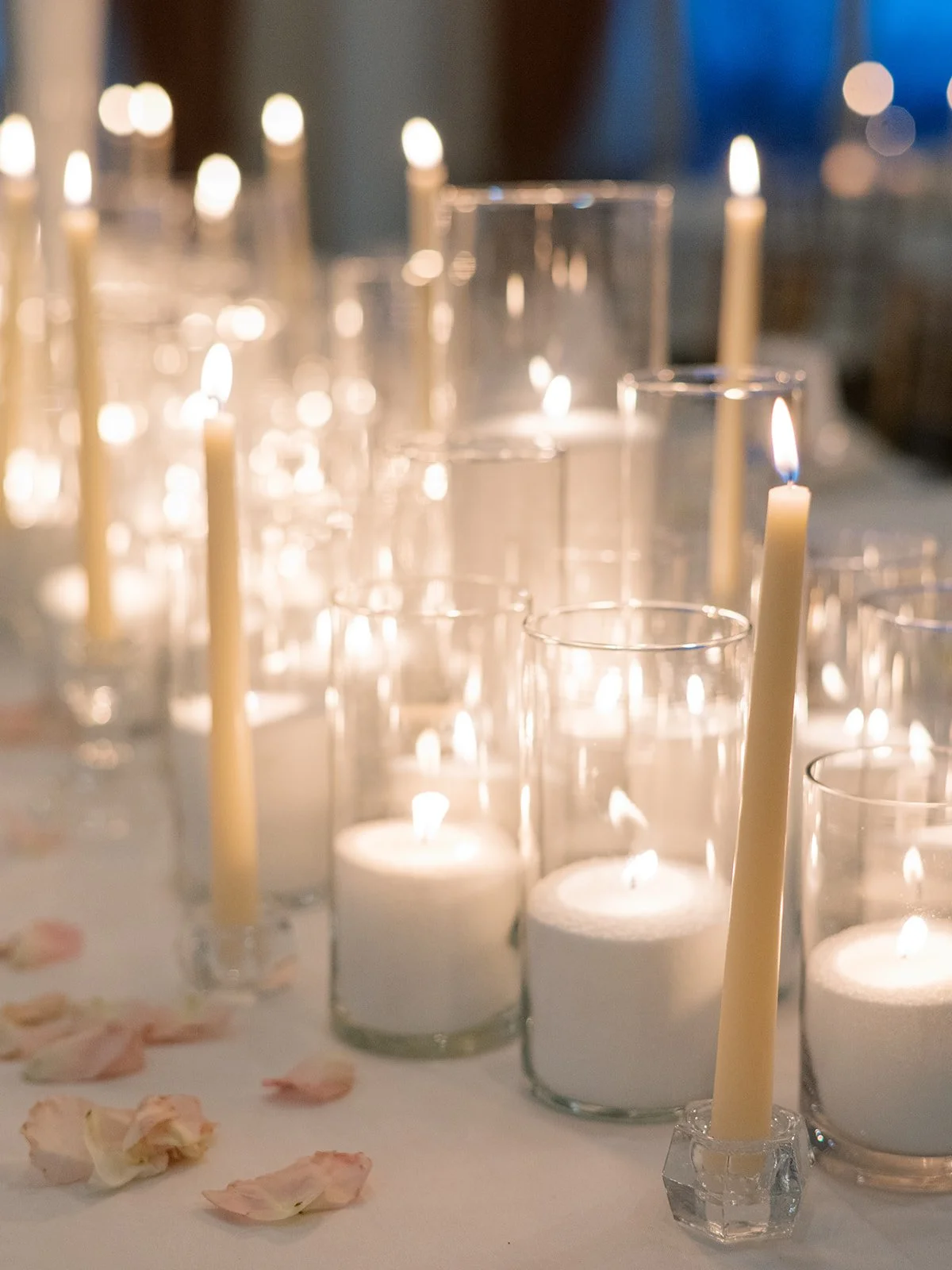 Multiple lit candles in glass holders on a table with scattered rose petals, creating a warm and romantic atmosphere.