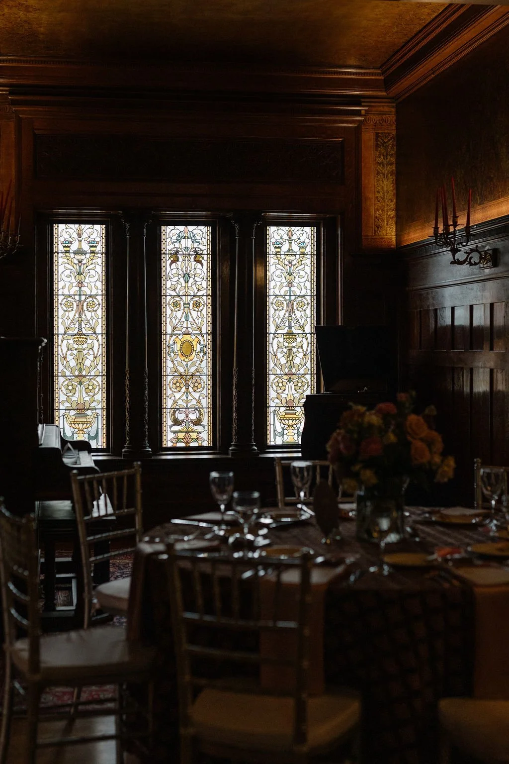 A dimly lit room with carved wooden walls and ceiling, featuring three stained glass windows with ornate patterns, a black grand piano, a chandelier with red candles, a table set with wine glasses and floral centerpiece, suggesting an elegant dining 