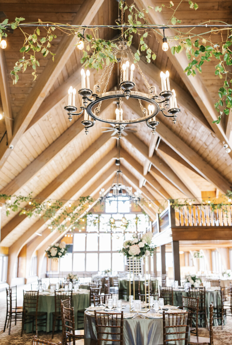 Elegant event space with high wooden vaulted ceilings, chandeliers, hanging string lights, and draped greenery, set up with round tables and chairs for a celebration.