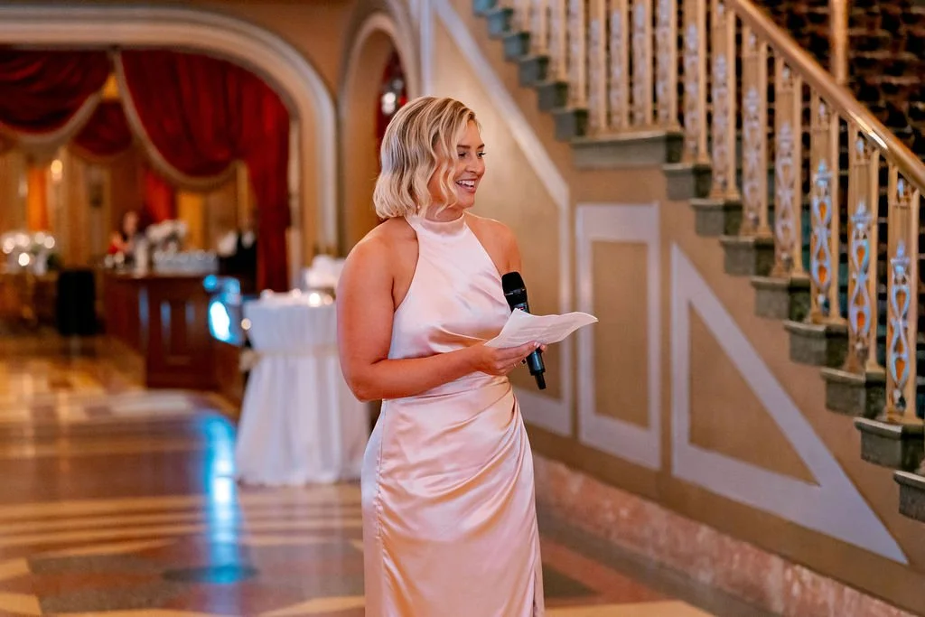 A woman in a satin dress holding a microphone and notes, smiling at an event in an elegant decorated hall with a staircase.