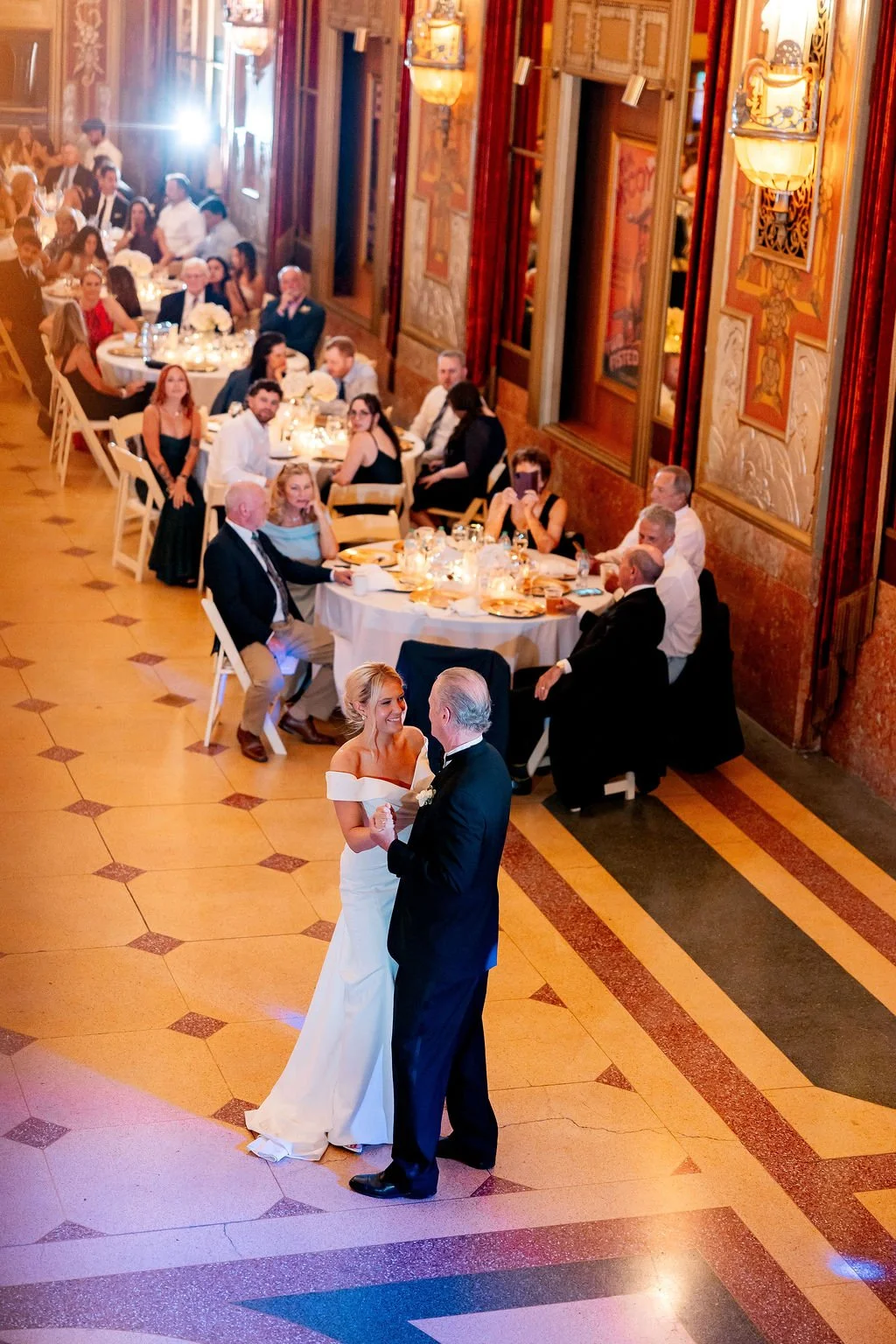 A bride and groom dancing at their wedding reception in a grand, ornate ballroom with guests seated at tables watching.