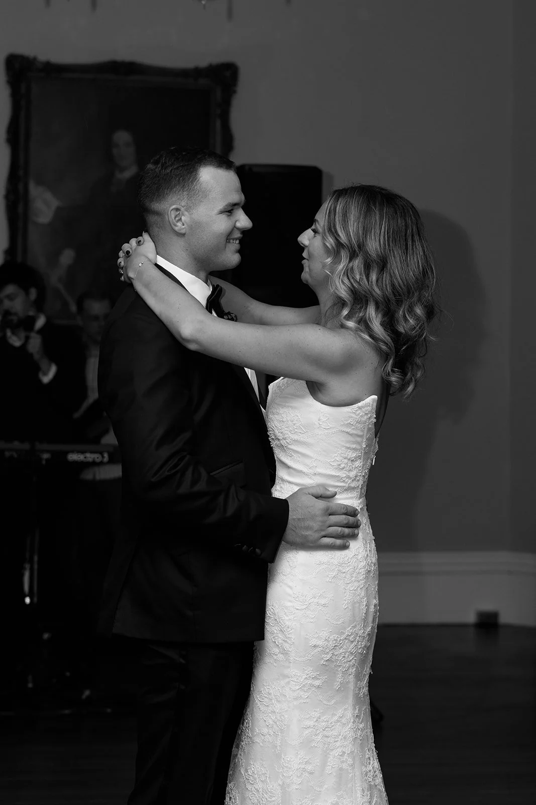 A black-and-white photo of a bride and groom dancing, looking at each other, with the bride smiling and the groom smiling. The bride is wearing a strapless lace wedding gown, and the groom is wearing a tuxedo with a bow tie. In the background, there 