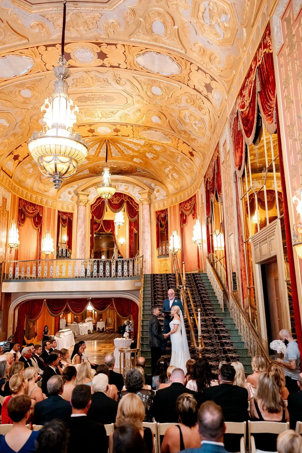 A wedding ceremony inside an ornate, grand hall with high, decorated ceilings, chandeliers, and red velvet drapes. The bride and groom are on the staircase, holding hands, with an officiant above them. Guests are seated facing the couple, and a photo