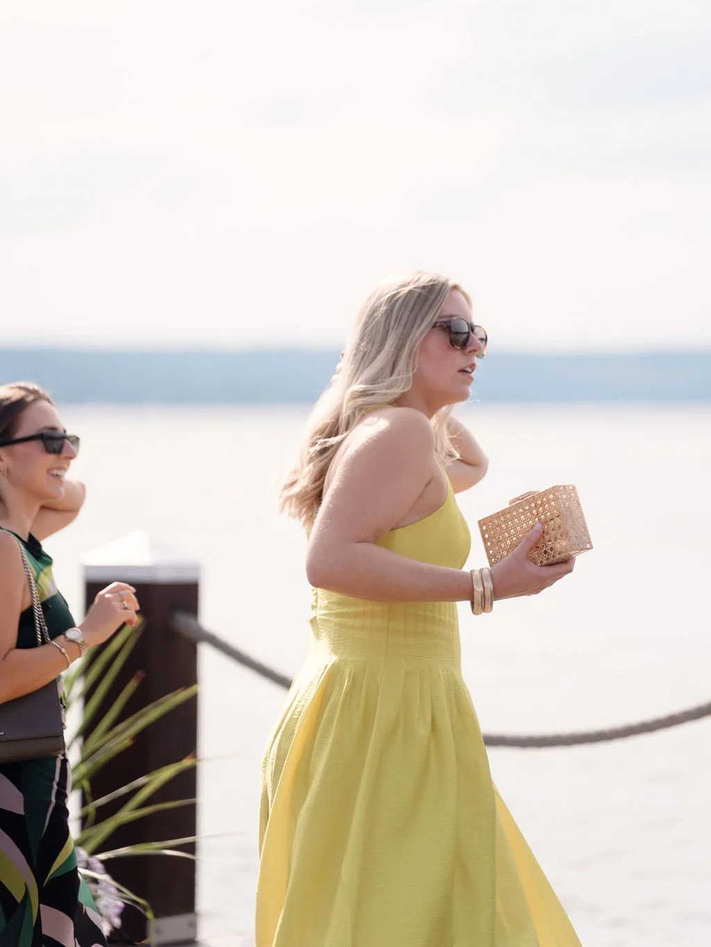 Two women enjoying a walk outdoors near water, one in a yellow dress holding a woven clutch, both wearing sunglasses.