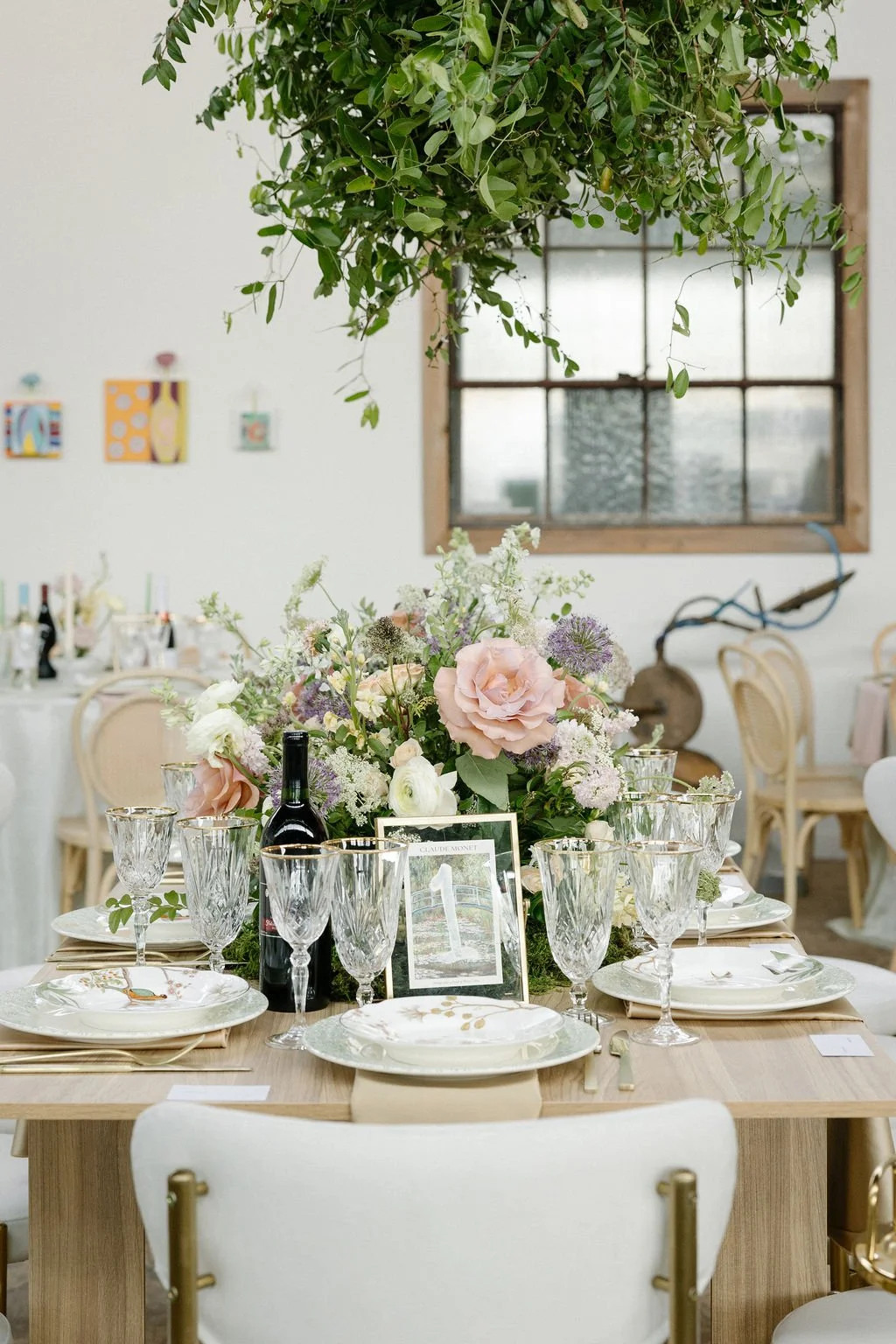 Elegant dining table set for a celebration with floral centerpiece, wine bottle, and glassware, in a room decorated with hanging greenery and modern art.