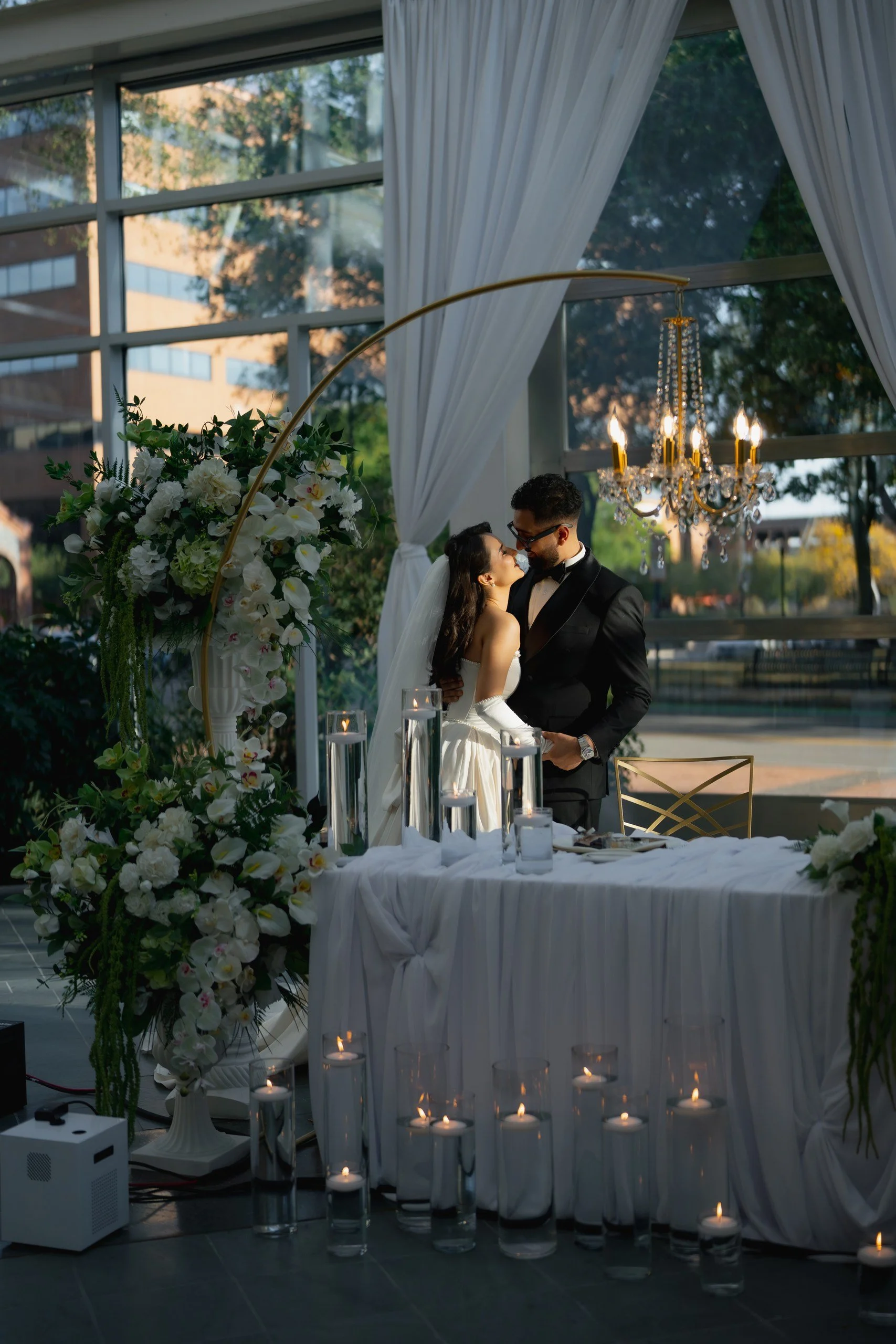 A wedding scene with a bride and groom near a table decorated with white flowers, candles, and a chandelier. The setting appears to be indoors with large windows and white curtains.