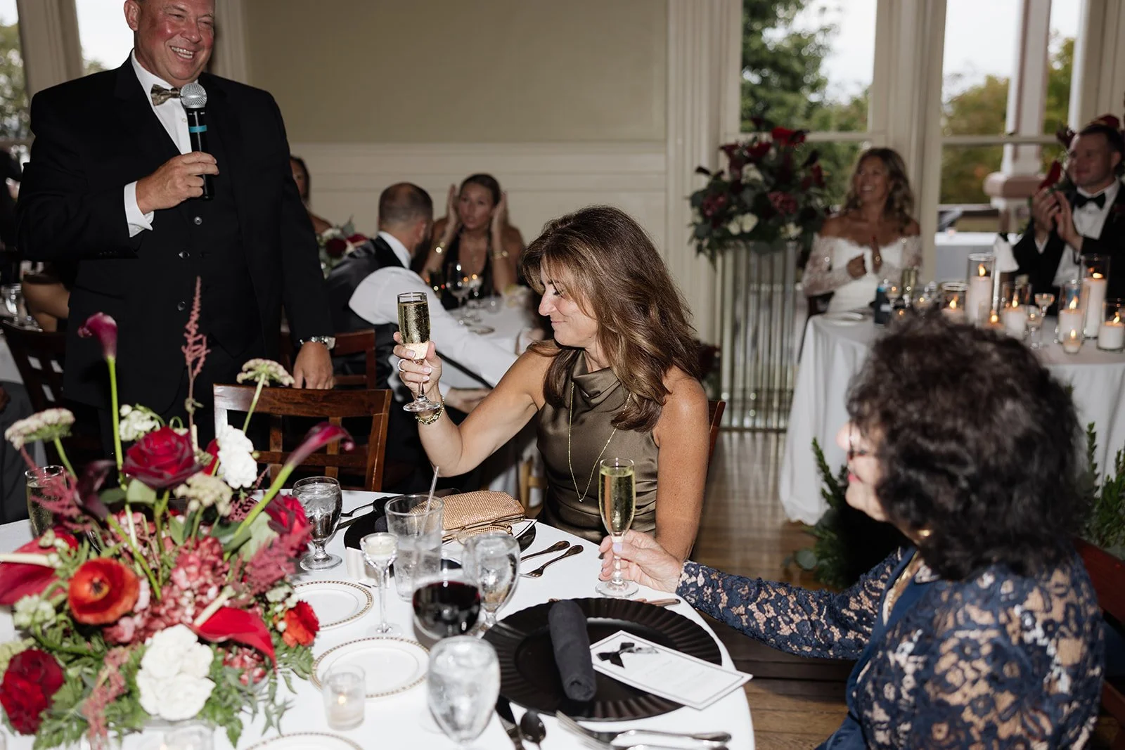 A man in a tuxedo giving a speech at a formal event, while women seated at a table raise their glasses in toast, surrounded by floral arrangements and elegant table settings.