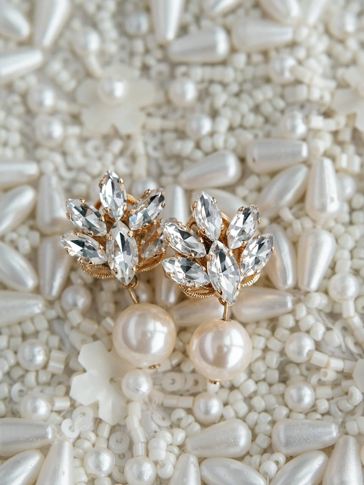 Elegant pearl earrings with large, clear, marquise-shaped rhinestones set in gold-tone metal, surrounded by small pearls, on a background of various white pearls and beads.