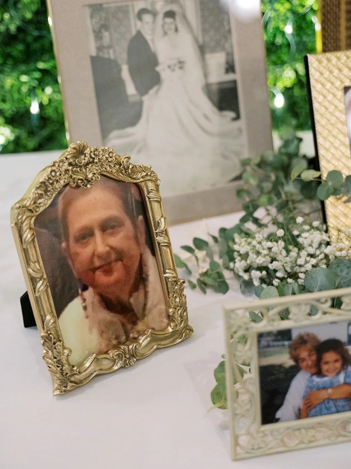 A collection of framed photographs and flowers arranged on a table, including a portrait of a smiling woman in a gold ornate frame, a black and white wedding photo in a gray frame, and a small picture of a woman and a child, with greenery in the back