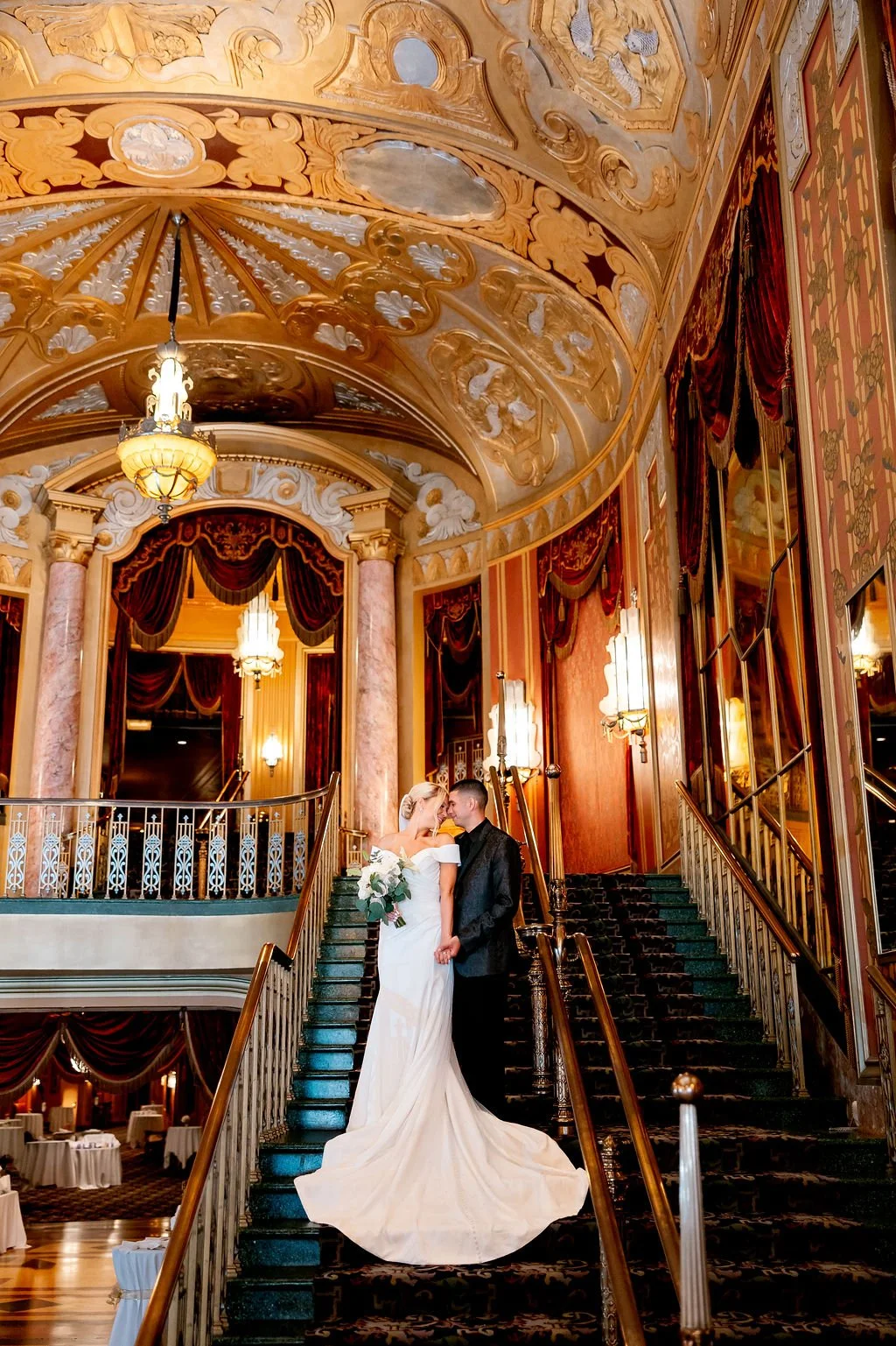 A bride and groom are standing on a grand staircase in an ornate, luxurious hall, sharing a romantic moment. The bride holds a bouquet and wears a white wedding gown with a train, while the groom wears a black tuxedo. The hall features elaborate gold