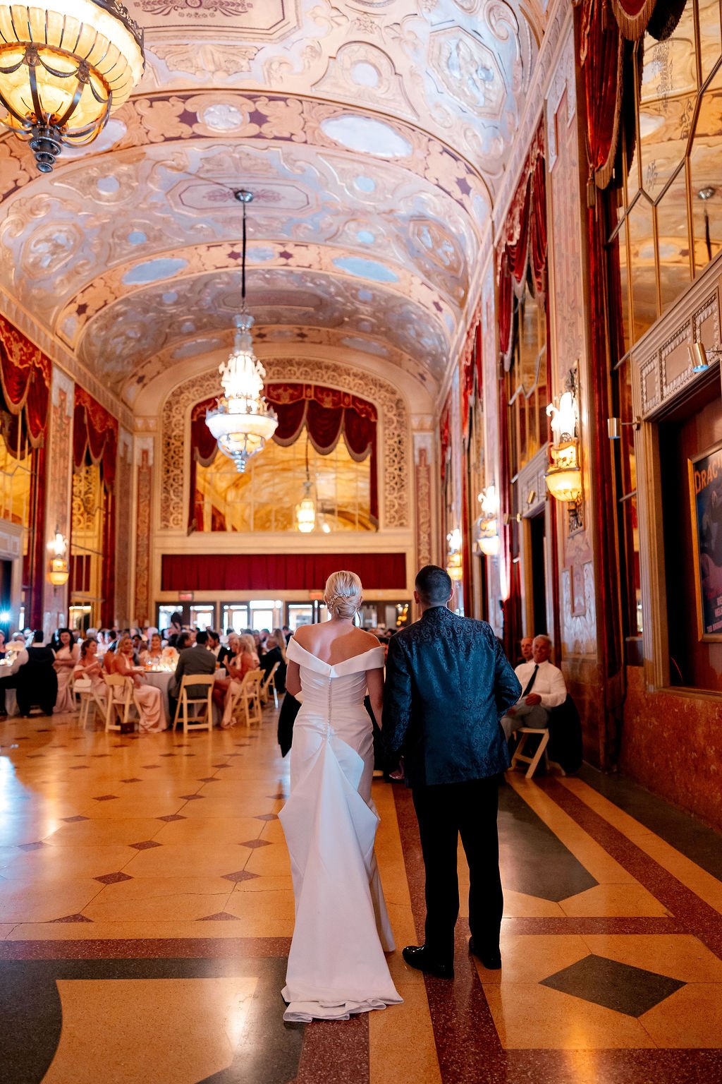 A bride and groom walking into a decorated banquet hall for their wedding reception, with guests seated at tables in the background.