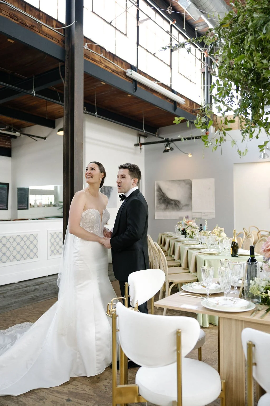 A bride and groom holding hands and smiling inside a decorated venue with long dining tables, wine bottles, and floral arrangements.