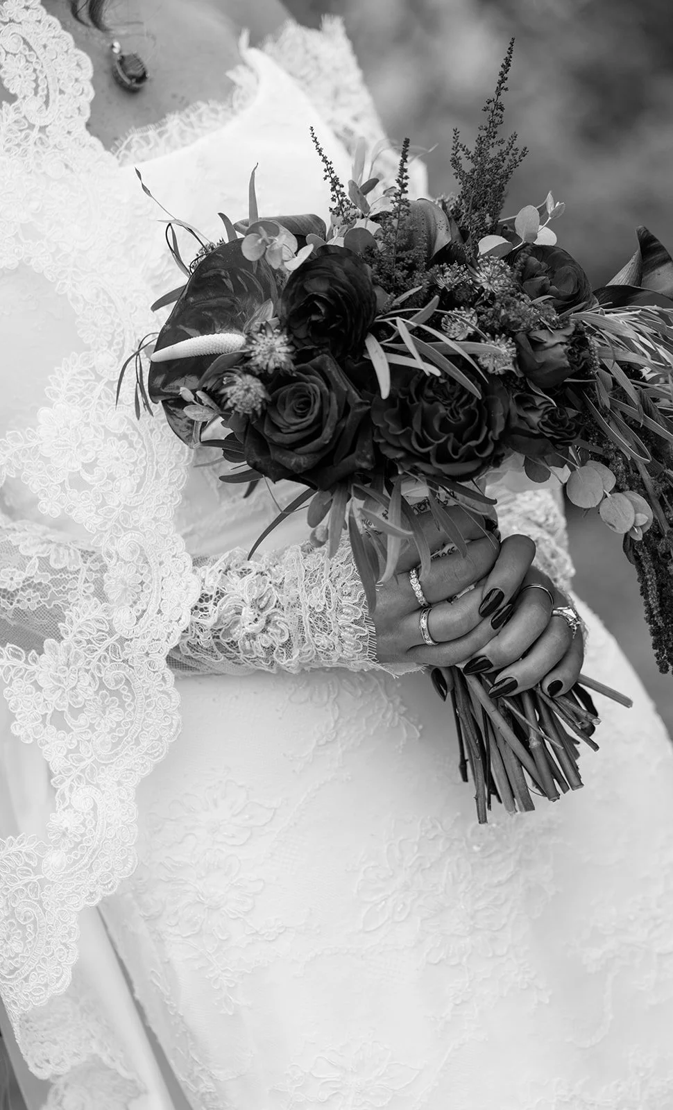 Close-up of a bride wearing lace wedding dress and holding a bouquet of flowers, with multiple rings on her fingers, in black and white.