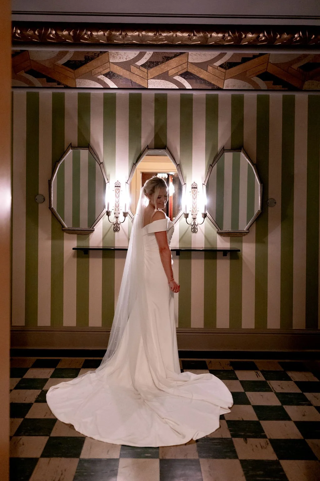 A bride in a white wedding dress with an off-shoulder design and a long train, standing in a decorative hallway with striped green and cream wallpaper, two octagonal mirrors, and wall sconces.