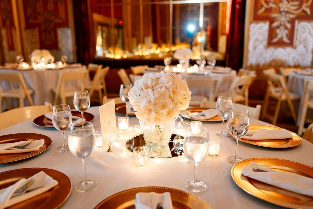 A round dining table set for a formal event with white flowers in a vase in the center, surrounded by glasses and place settings with gold chargers and white napkins, in an ornately decorated room with warm lighting.