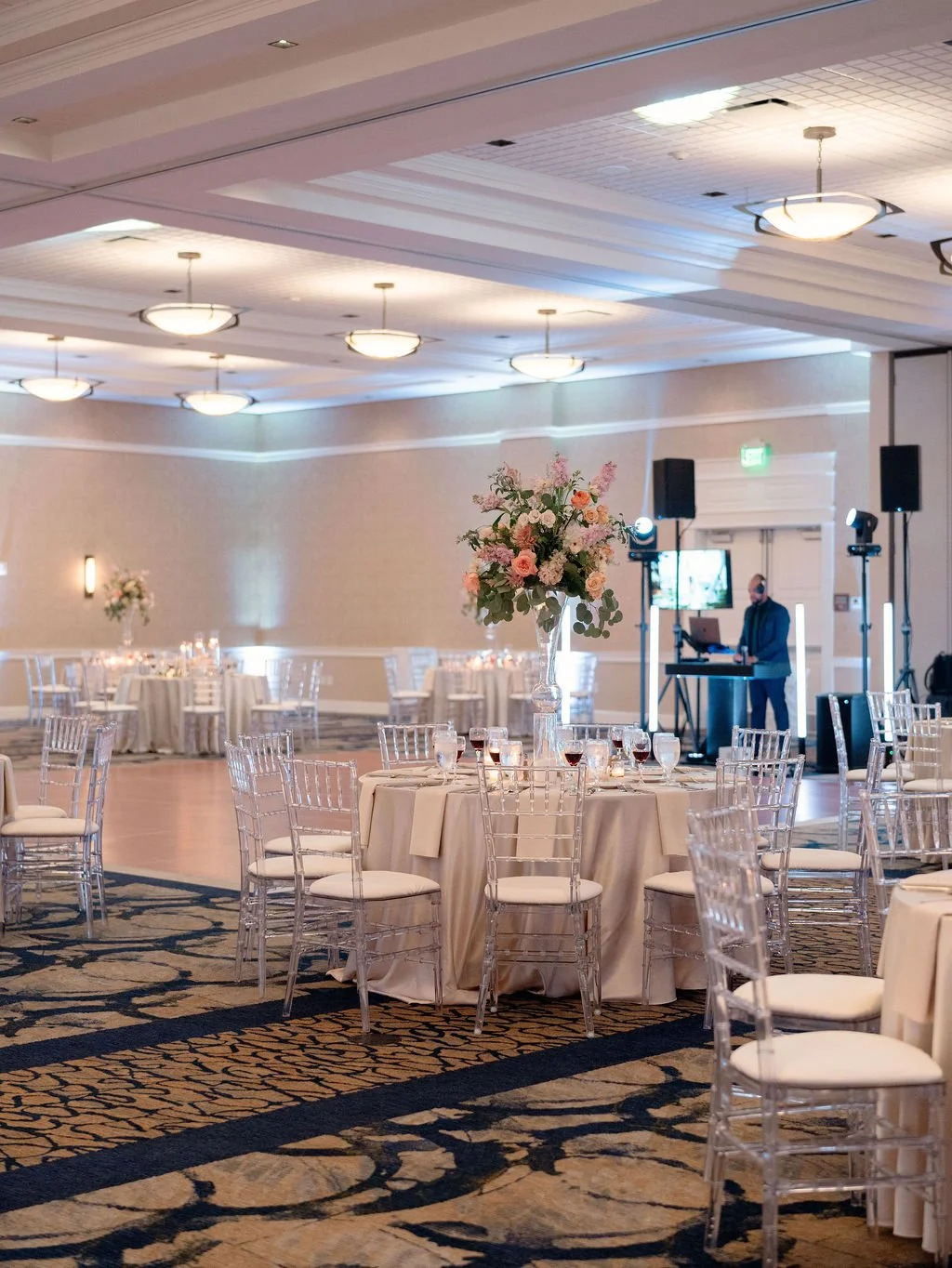 Elegant banquet hall decorated with flower arrangements, round tables with white tablecloths, and clear chairs. A large floral centerpiece is on a main table, and a DJ setup with lighting is present in the background.