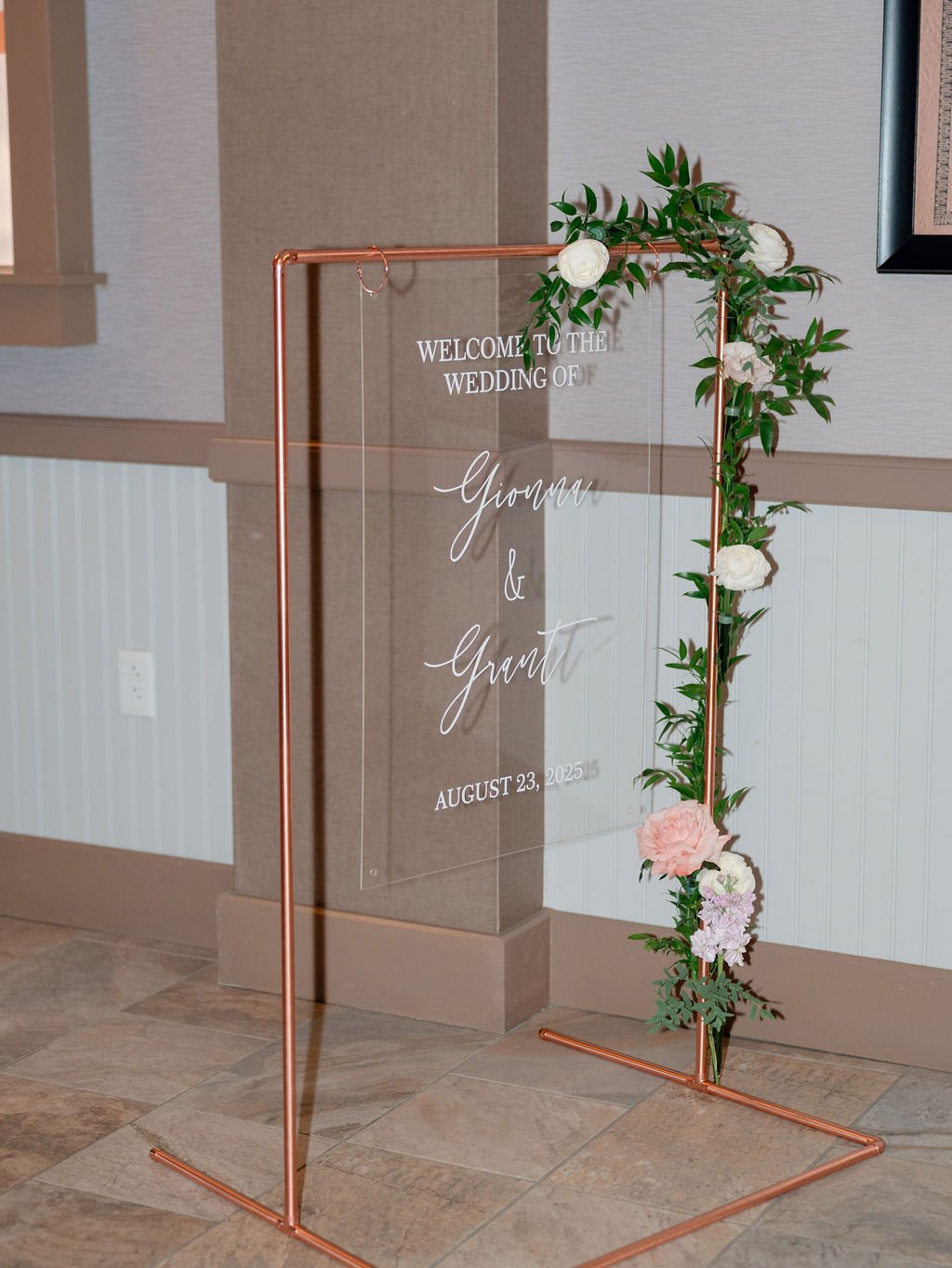 A decorative wedding welcome sign made of a clear acrylic panel with white cursive text, framed by a copper stand and adorned with artificial flowers and greenery on the right side.