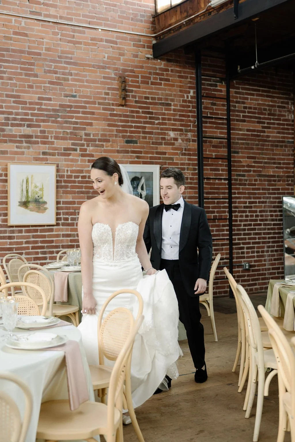 A bride in a wedding dress and a groom in a tuxedo walking in a restaurant with brick walls and wooden floors.