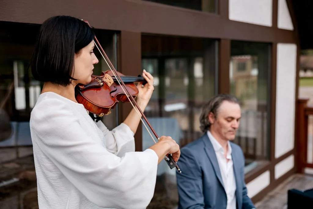 A woman playing the violin outdoors with a man sitting nearby and a wooden building in the background.