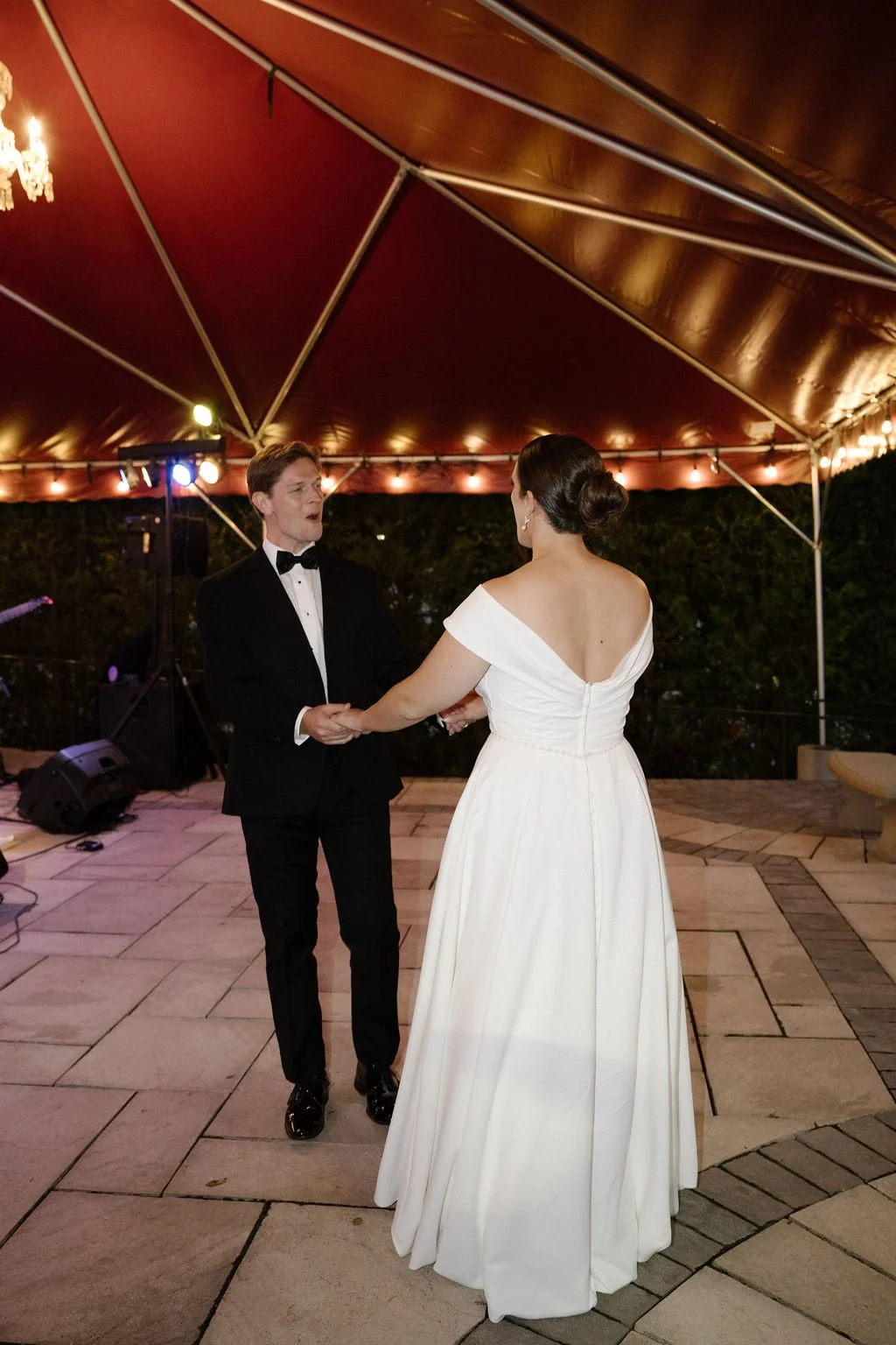 A bride and groom are dancing at their wedding reception under a large outdoor tent at night, with decorative lighting and a band or music setup nearby.