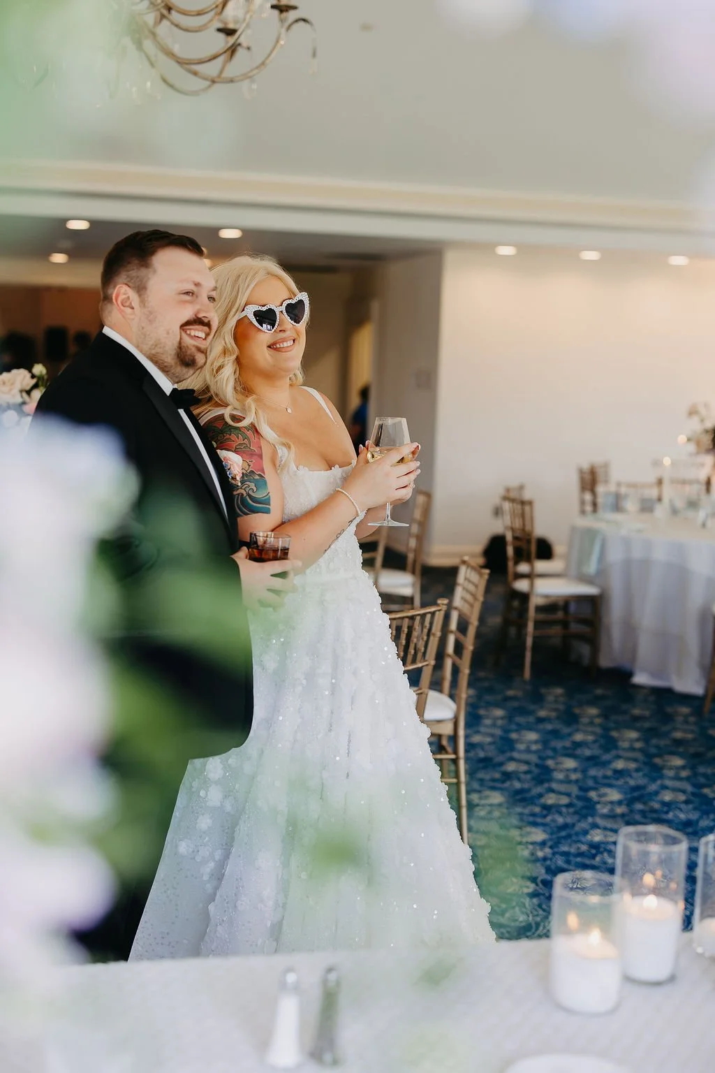A bride and groom standing together at their wedding reception, both holding drinks and smiling, with the bride wearing heart-shaped sunglasses.