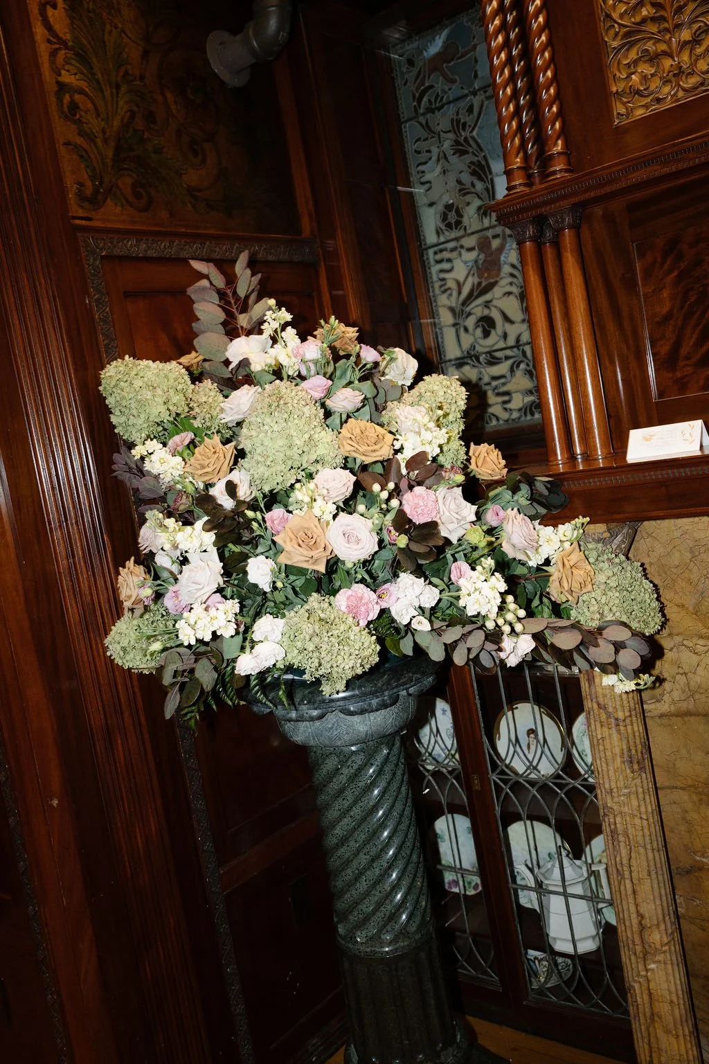 A large floral arrangement with roses, hydrangeas, and other flowers in a tall, dark, spiral column vase, set against polished wood and glass display cabinets with decorative wood detailing.