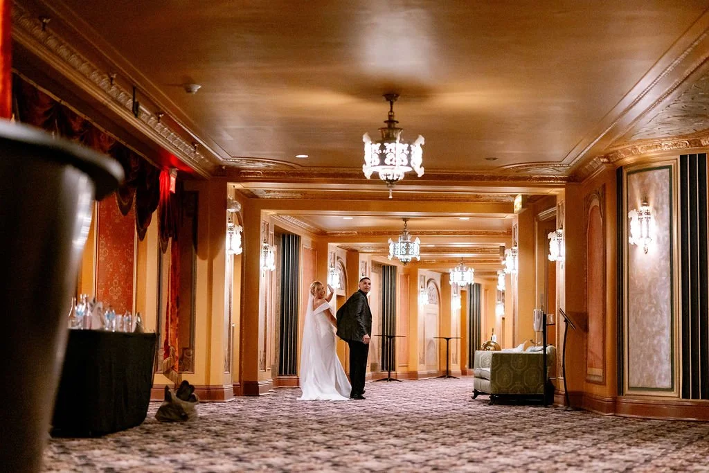 A bride in a white wedding gown and a groom in a dark suit standing in a luxurious hotel corridor with ornate chandeliers and elegant decor.