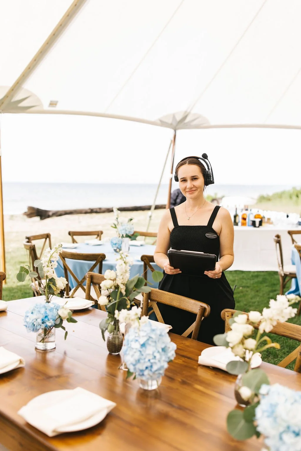 A woman standing at a decorated outdoor event table holding a tablet, wearing a headset and a black dress.