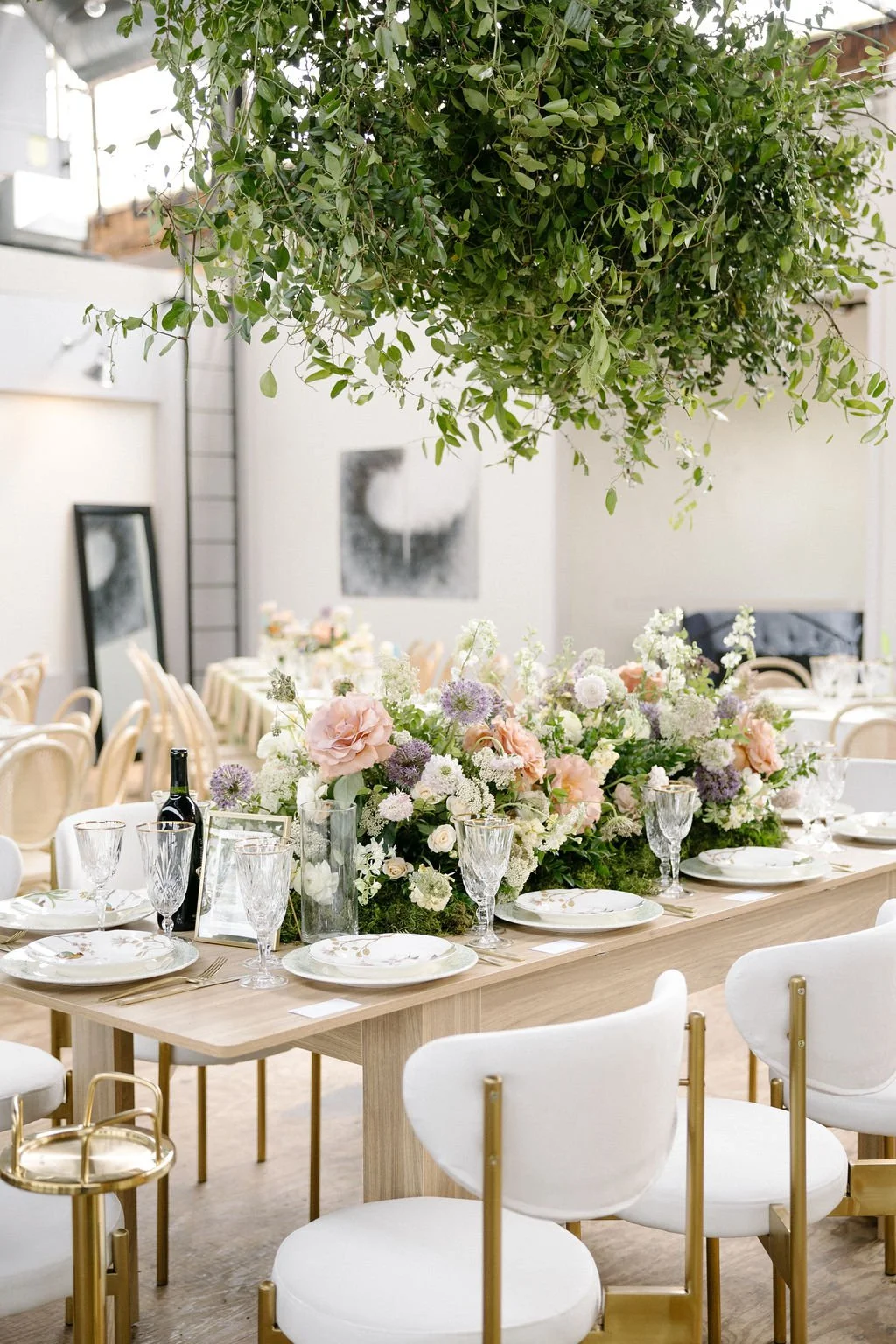 Elegant dining table set with floral centerpieces in a bright, modern space, featuring hanging greenery and artwork.