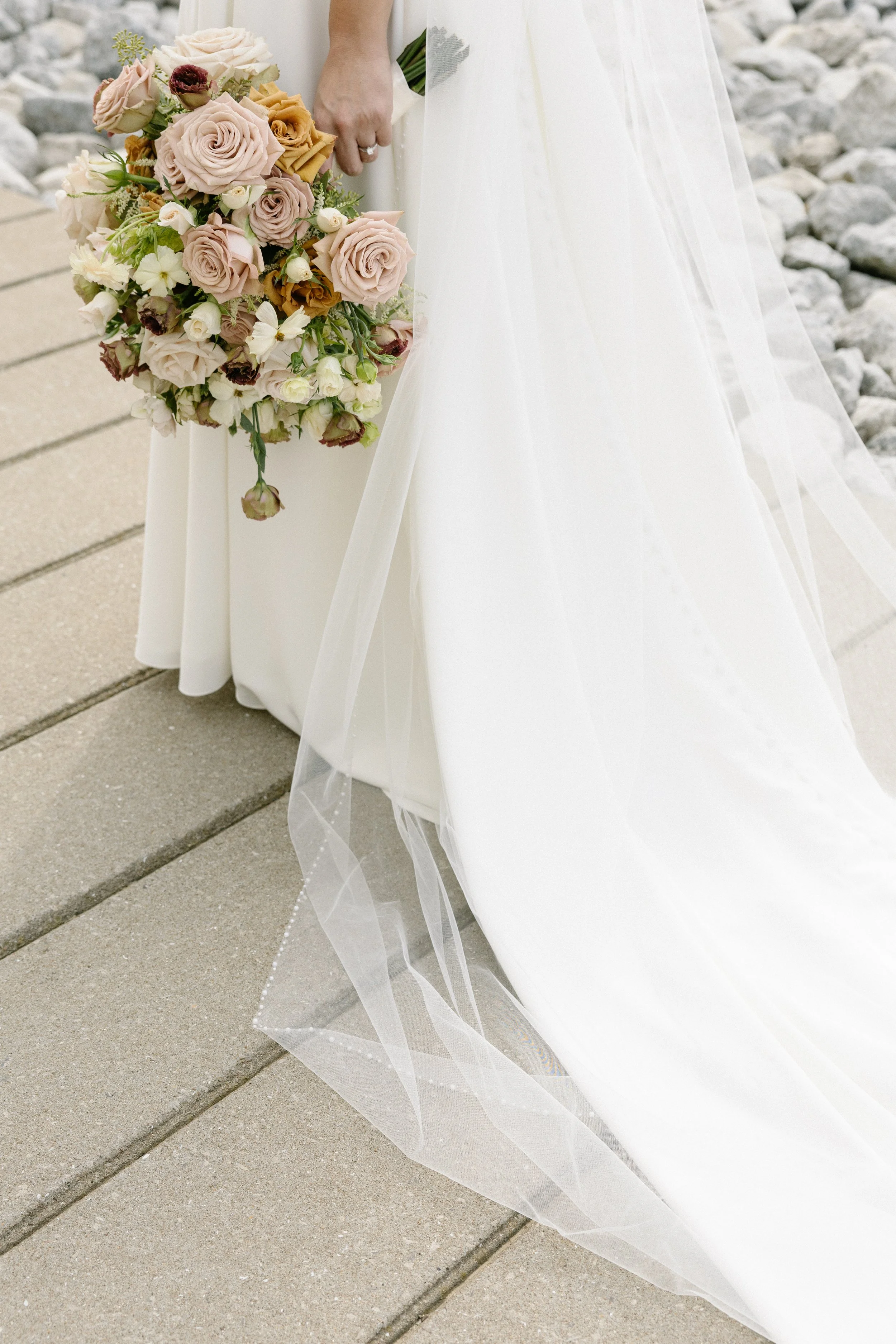 Bride holding a cascading bouquet of beige, cream, and burgundy flowers, standing on a sidewalk with rocks in the background.