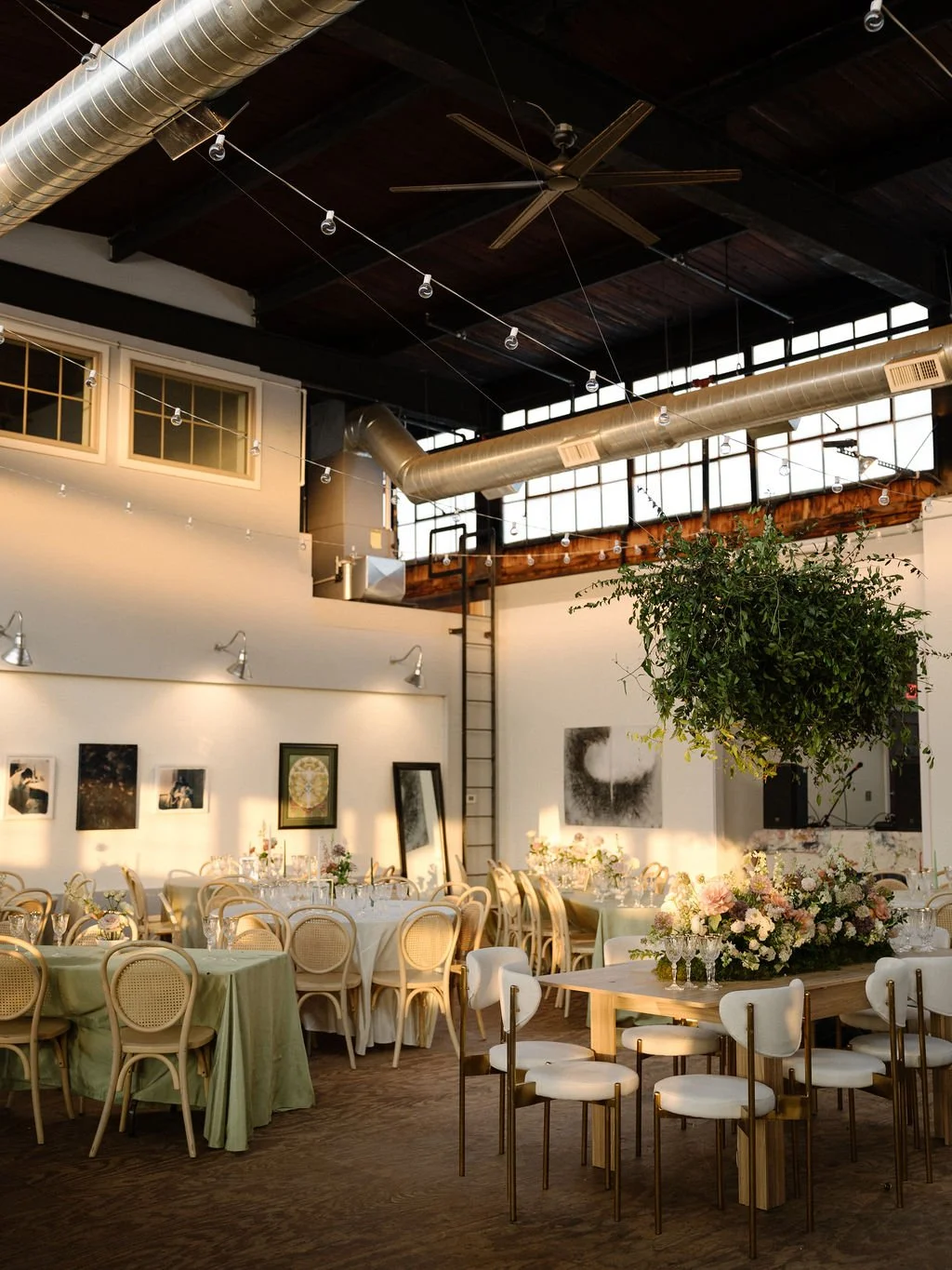 Interior of a decorated event space with round tables covered in pastel tablecloths, floral centerpieces, and chairs with woven backs. A large hanging plant is above, with artwork on the walls and string lights across the ceiling, and large industria