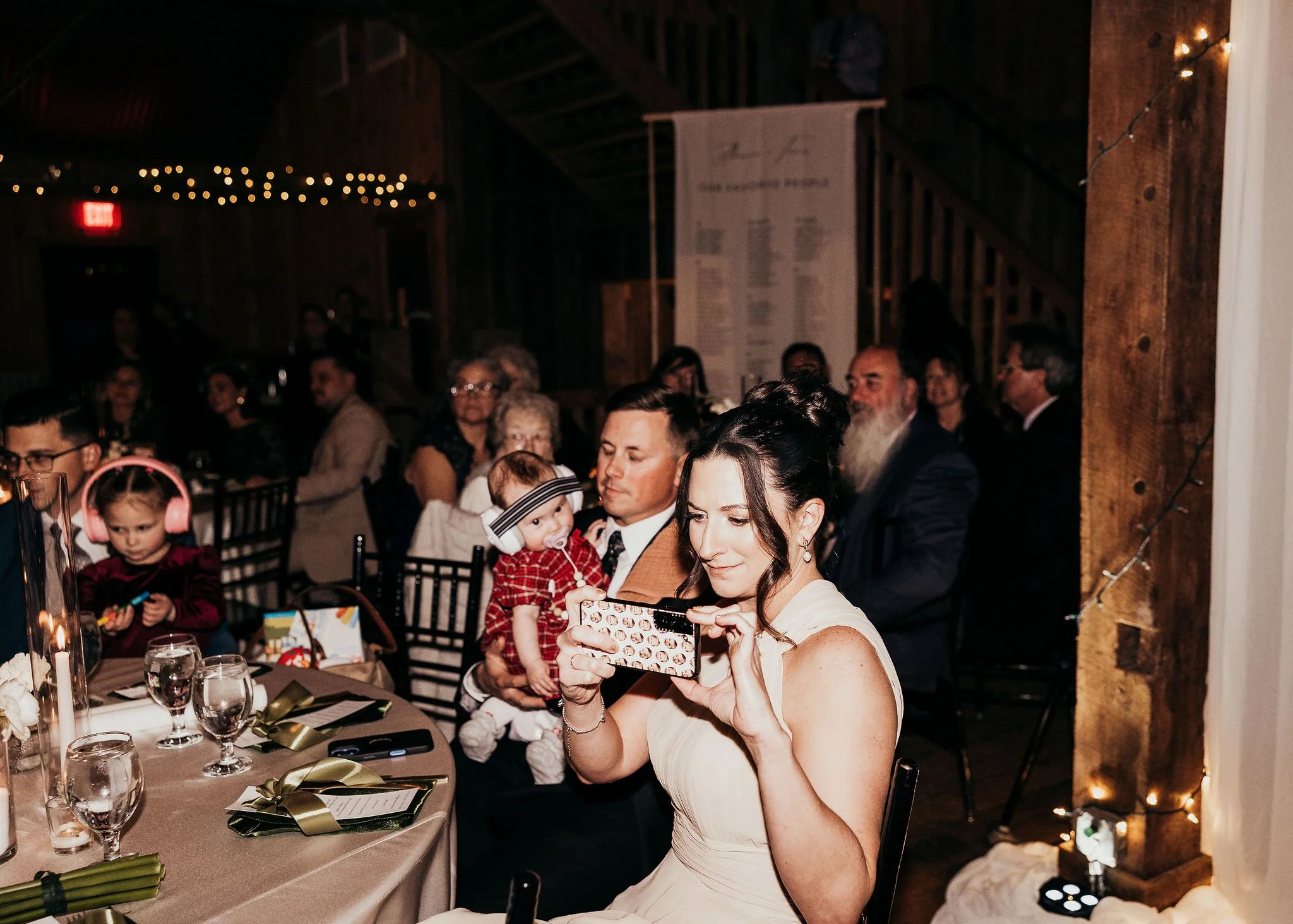 A woman in a white dress takes a photo with her phone at a festive gathering with children and adults seated at tables, dark wood interior with string lights and an informational poster in the background.